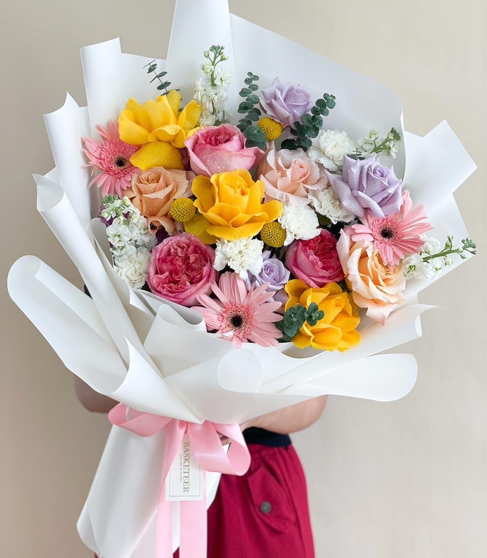 Vibrant pastel rose and gerbera bouquet with yellow, pink, peach, and purple blooms, white-wrapped with pink ribbon