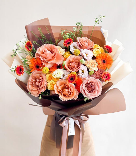Sunset-toned fresh flower bouquet with peach roses, orange gerberas, and yellow craspedia in luxury brown paper wrap