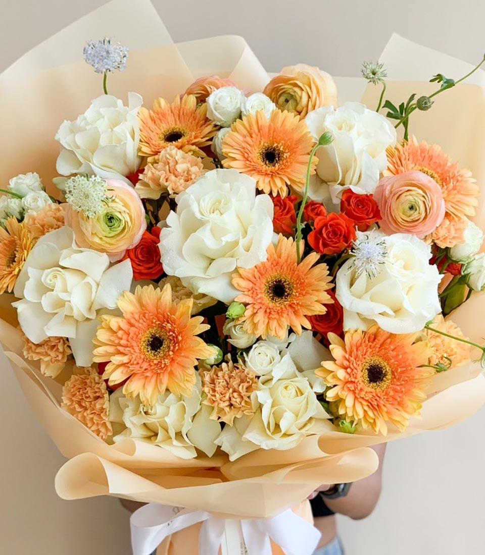 Warm peach and cream rose bouquet with orange gerbera daisies and ranunculus, wrapped in beige paper with white ribbon