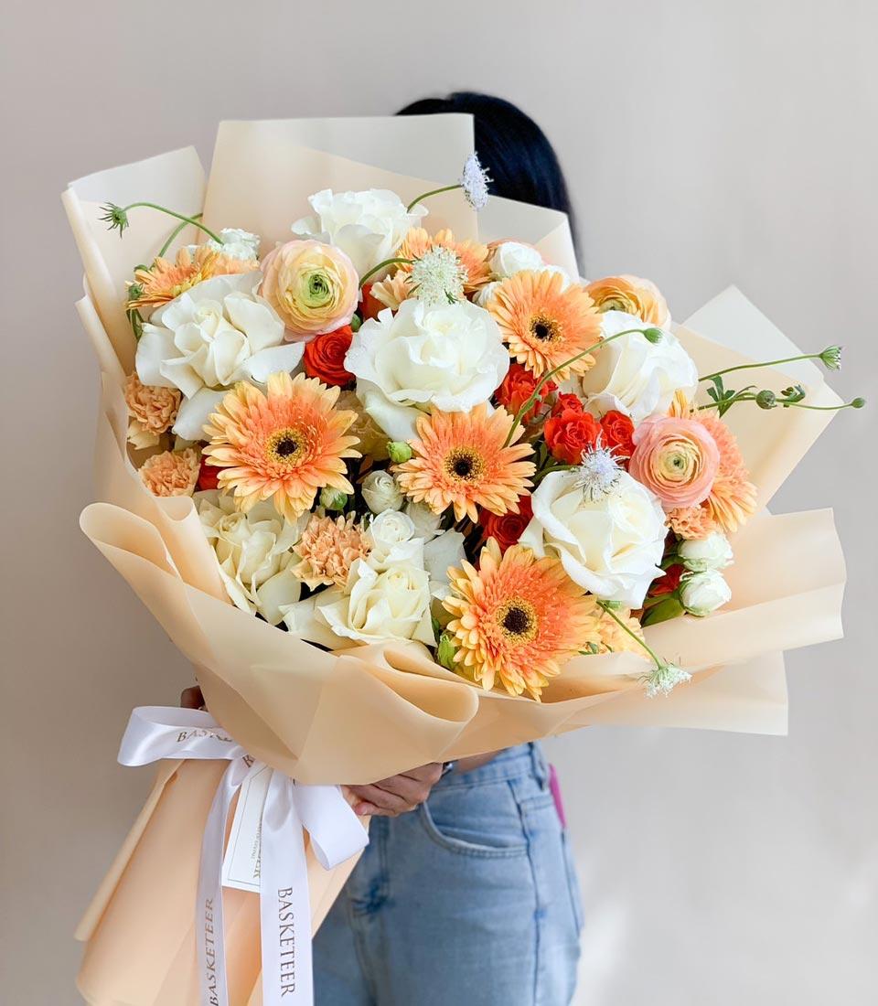 Warm peach radiance bouquet with orange gerberas, cream roses, and ranunculus wrapped in peachy paper with white ribbon