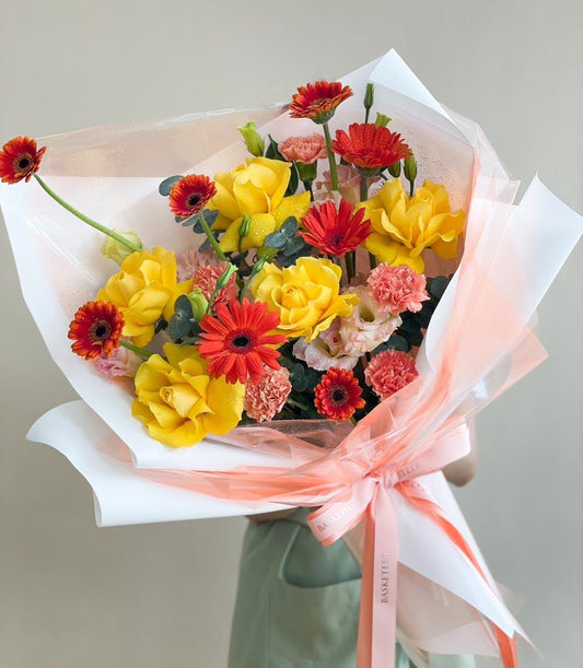 Vibrant yellow roses and red gerberas with pink carnations, wrapped in peach paper and satin ribbon