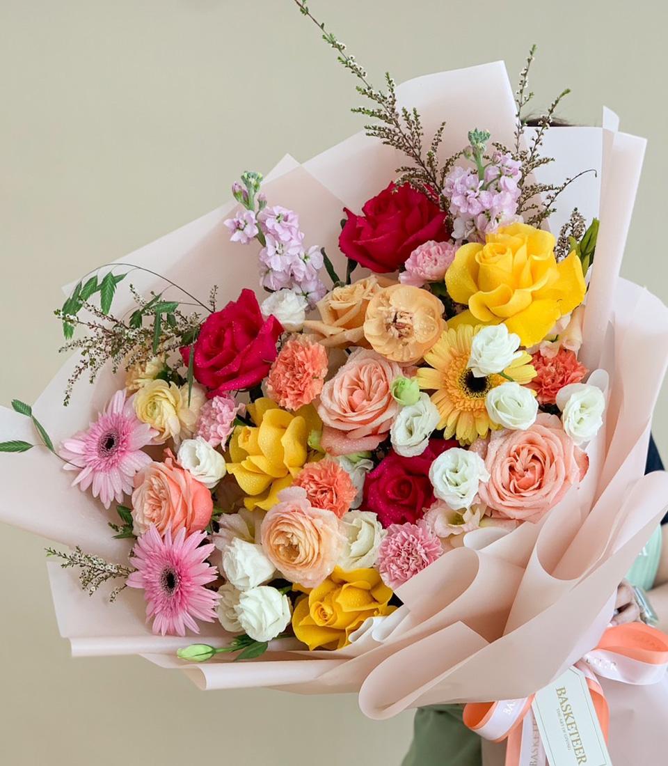 Vibrant mixed rose and gerbera bouquet with red, yellow, peach, and pink blooms in light pink wrapping