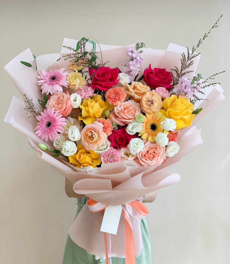 Vibrant mixed flower bouquet with pink gerberas, red and yellow roses, orange carnations, and purple stock in light pink