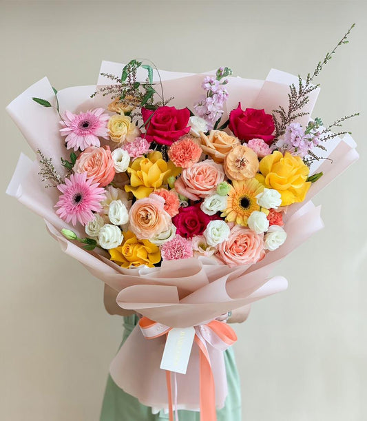 Vibrant mixed flower bouquet with pink gerberas, red and yellow roses, orange carnations, and purple stock in light pink