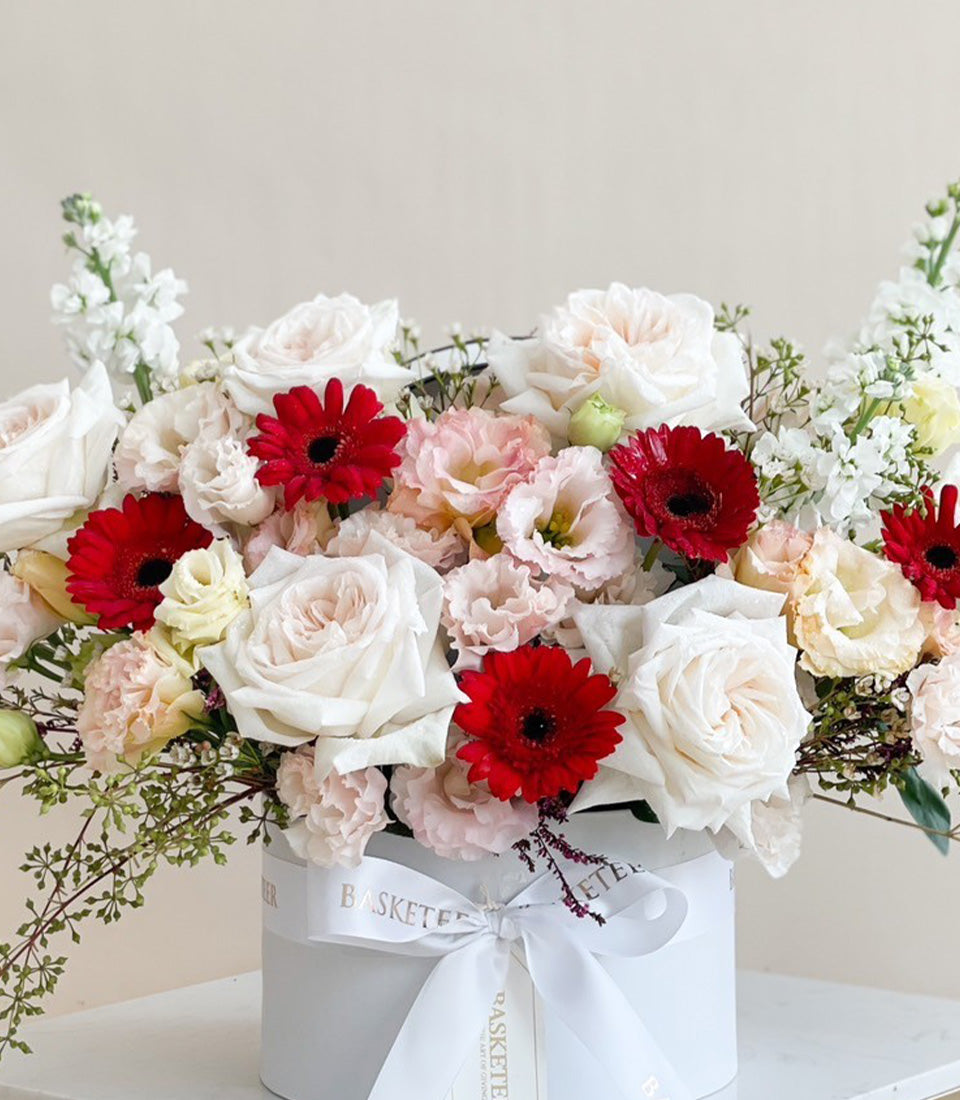 Luxe white cylindrical box with white and red roses, gerbera daisies, and lisianthus, finished with satin ribbon bow
