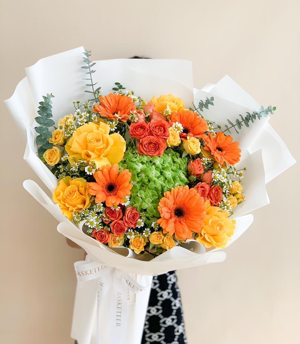 Vibrant orange and yellow rose bouquet with gerbera daisies, hydrangea, and eucalyptus in white paper wrapping