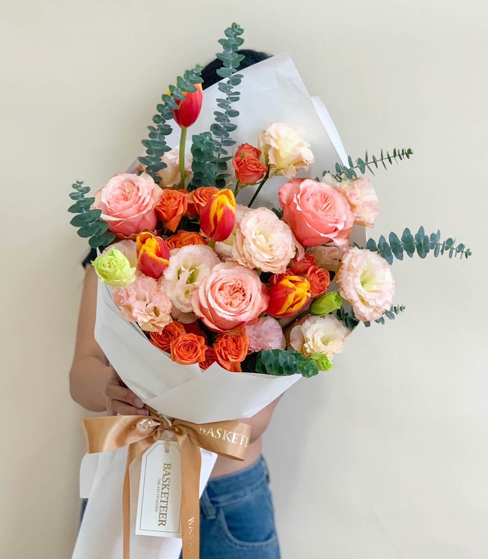 Vibrant mixed bouquet of pink and orange roses, red and yellow tulips, lisianthus with eucalyptus, wrapped in white paper