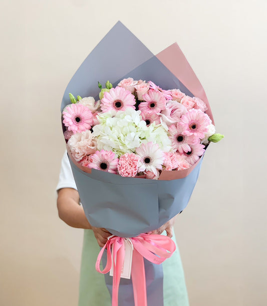Fresh pink gerbera daisies, white hydrangeas, and roses wrapped in grey-blue and blush paper with pink ribbon bow