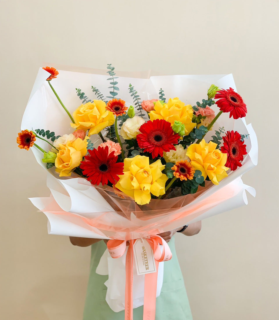 Vibrant bouquet of red gerberas, yellow roses, and pink carnations with eucalyptus, wrapped in peach paper