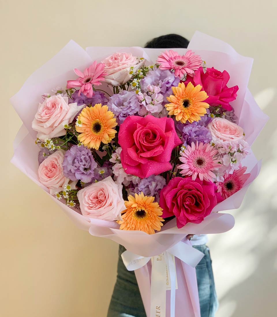 Vibrant mixed bouquet of hot pink roses, orange gerbera daisies, and purple eustoma with fresh water droplets, wrapped in