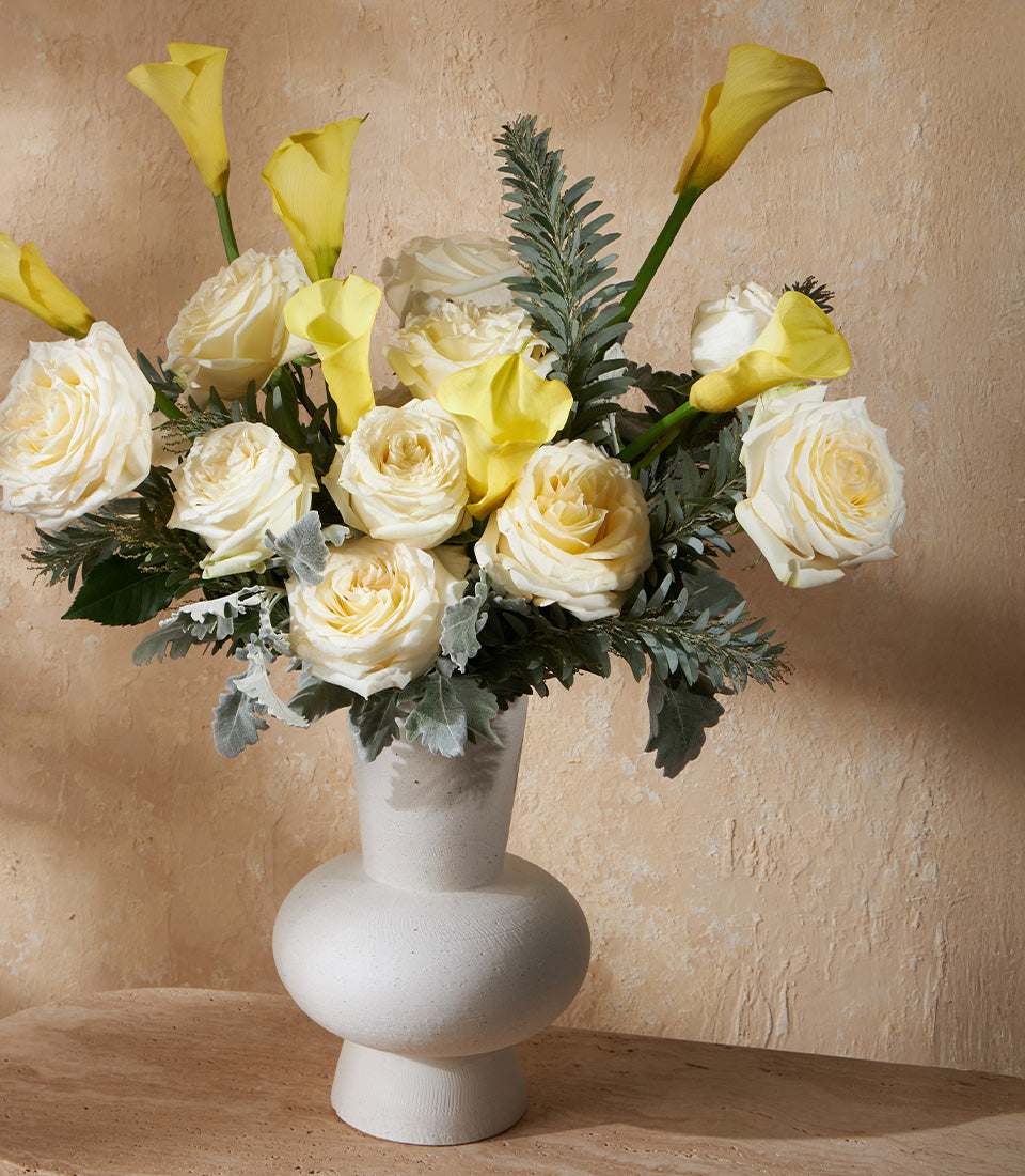 Cream white roses and yellow calla lilies in textured ceramic vase with green foliage