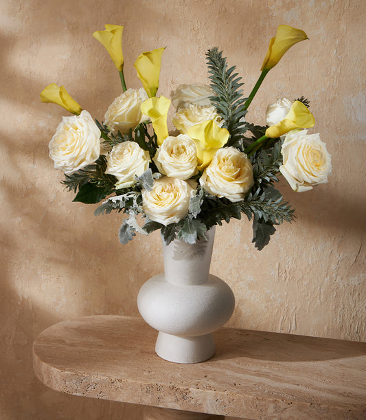 Yellow and white flower arrangement in white textured vase with roses, calla lilies, and greenery