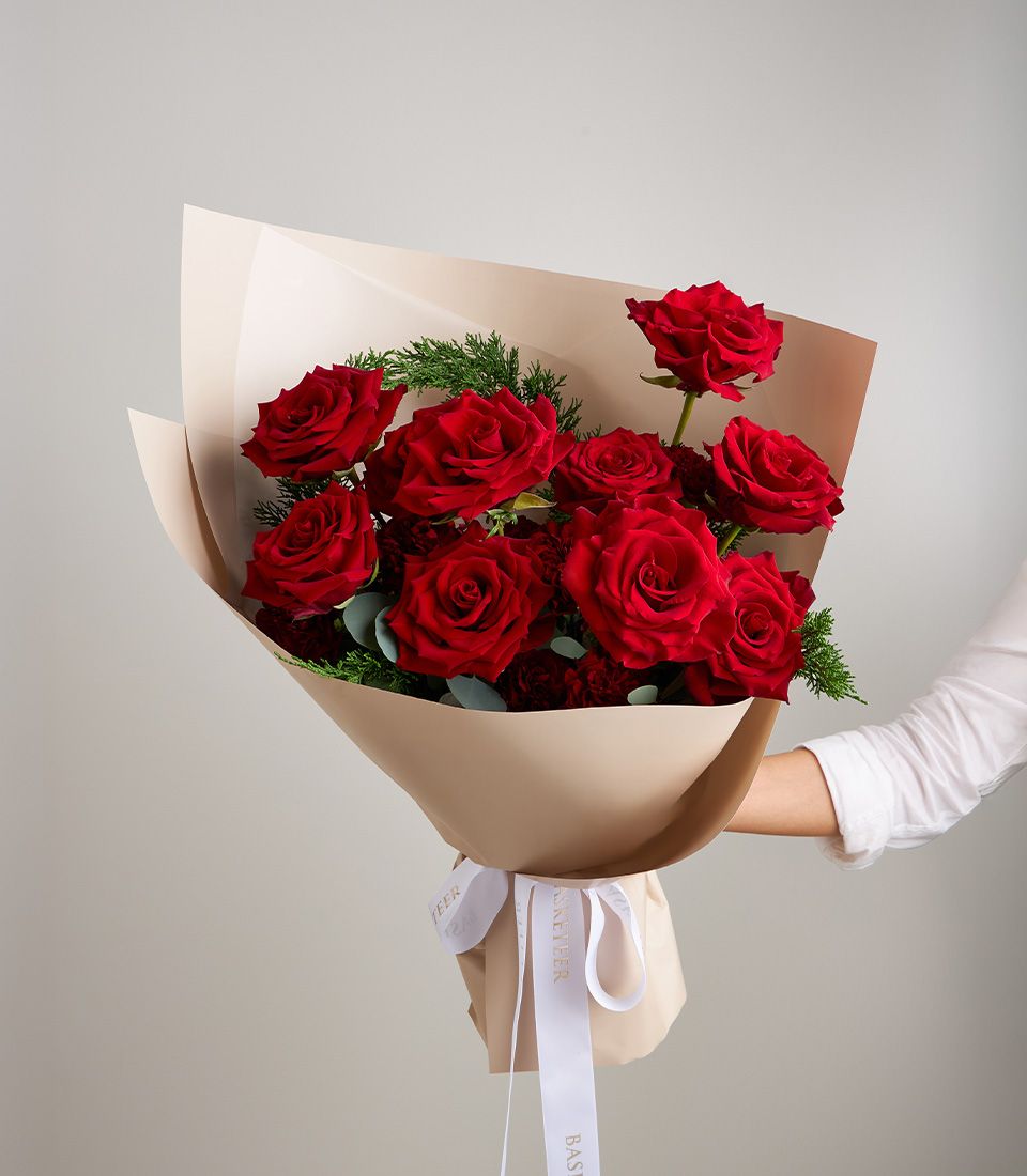 Vibrant red rose bouquet with green foliage, wrapped in kraft paper with white ribbon, held by hand against grey background