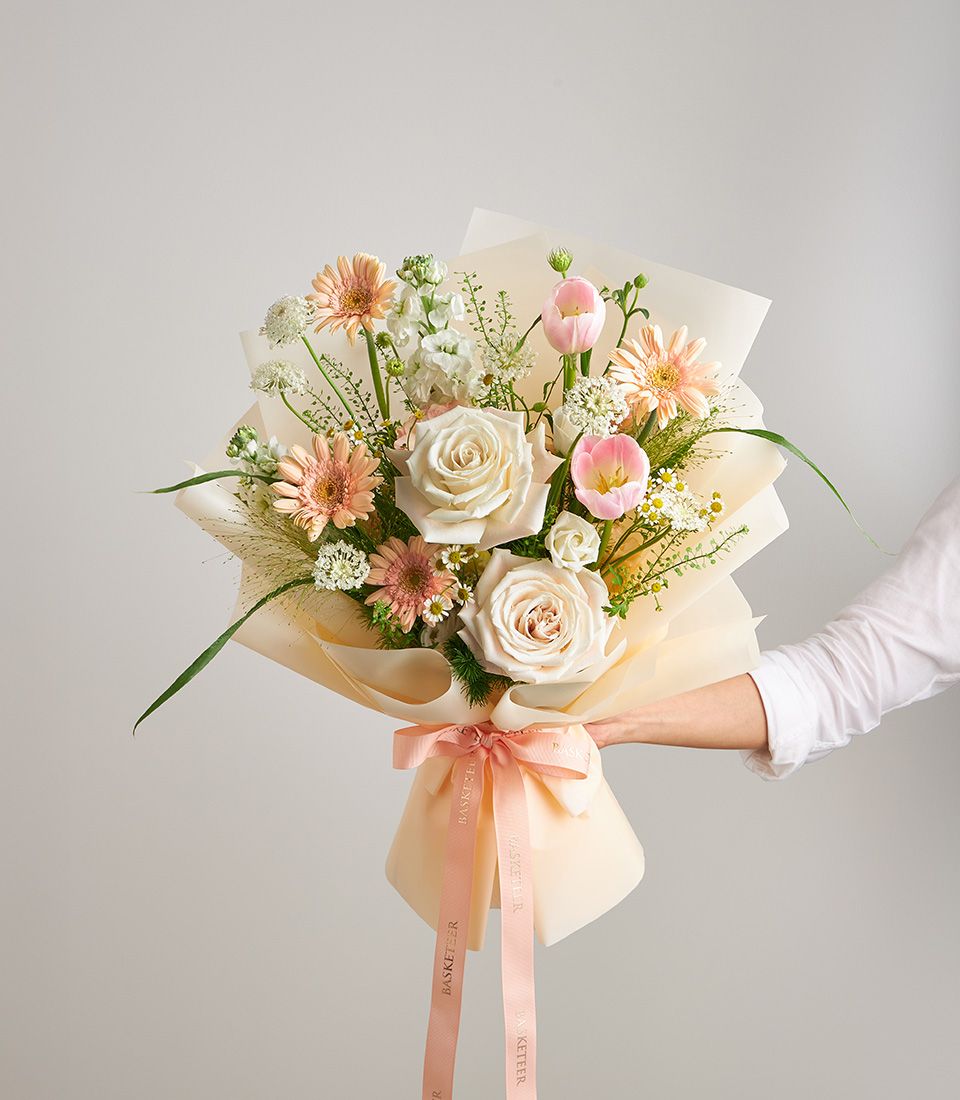Hand-held mixed bloom bouquet with cream roses, pink gerberas, and tulips in peach ribbon wrapping