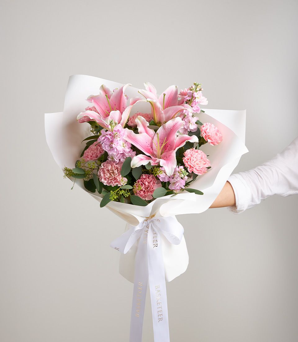 Romantic pink lily bouquet with speckled petals, carnations, and purple stock wrapped in white paper with gold ribbon bow