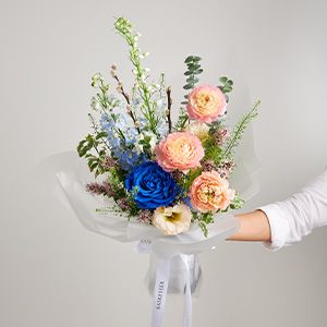 Hand holding blue rose and peach pink bouquet wrapped in white paper with white ribbon bow