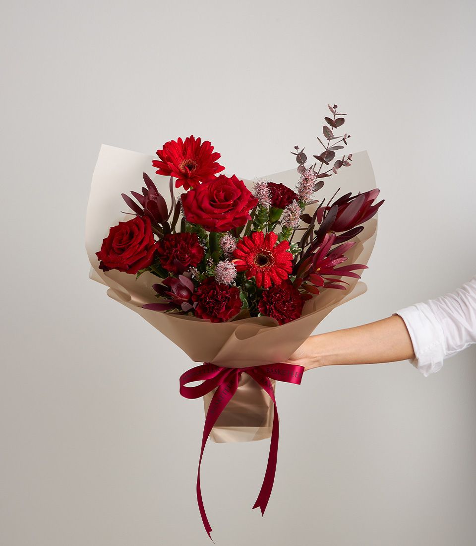 Hand holding passionate red mixed flower bouquet with roses, gerberas, and carnations in kraft paper with red ribbon