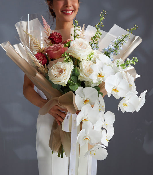 Luxury rose and orchid bouquet with white roses, pink astilbe, red dahlias, and blue delphiniums in beige paper