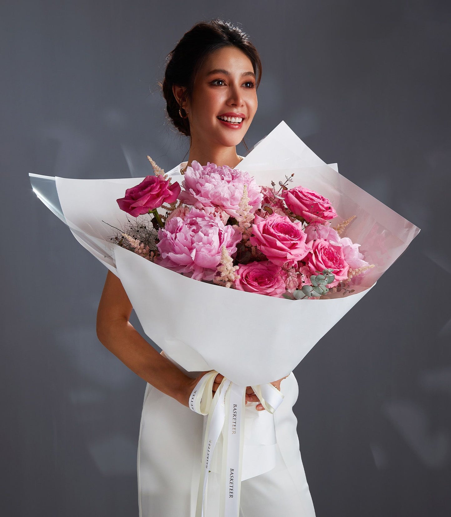 Woman holding luxury pink peony and rose bouquet with white ribbon wrapping and green foliage