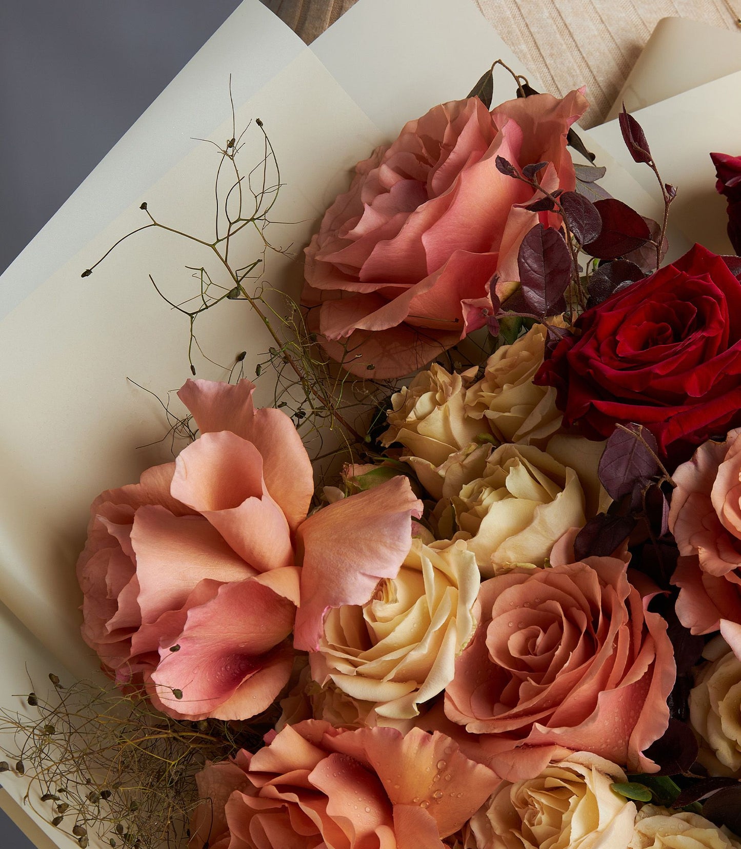 Fresh Ecuador roses in peach, coral, cream, and deep red with textured greenery and dried botanicals, wrapped in soft paper