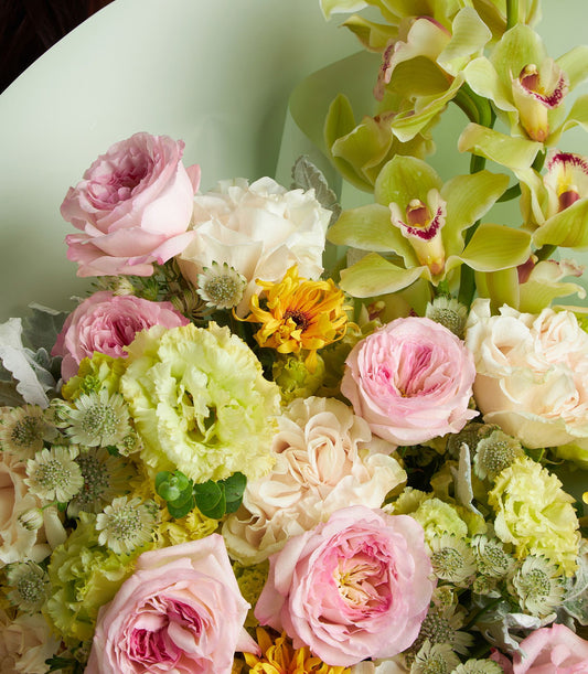Pastel mixed flower bouquet with pink roses, white lisianthus, yellow mums, and chartreuse orchids in light green wrap