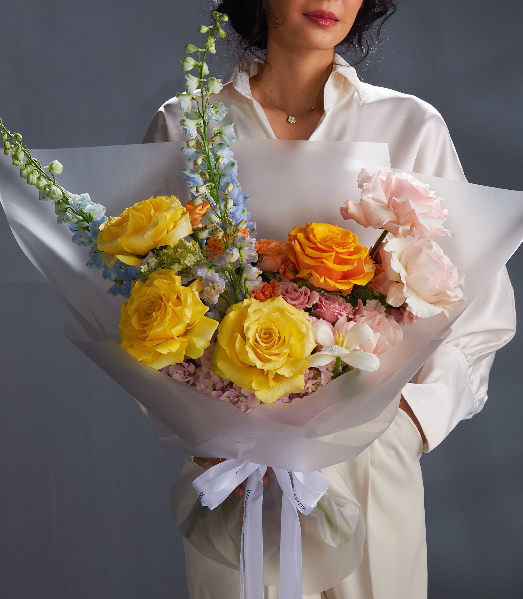 Radiant Bloom rose bouquet with yellow, orange, and pink roses, delphiniums, and accent flowers by Basketeer