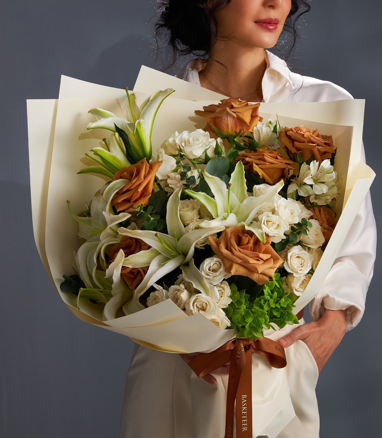Luxury lily and rose bouquet with white lilies, rust roses, and cream paper wrapping tied with brown satin ribbon