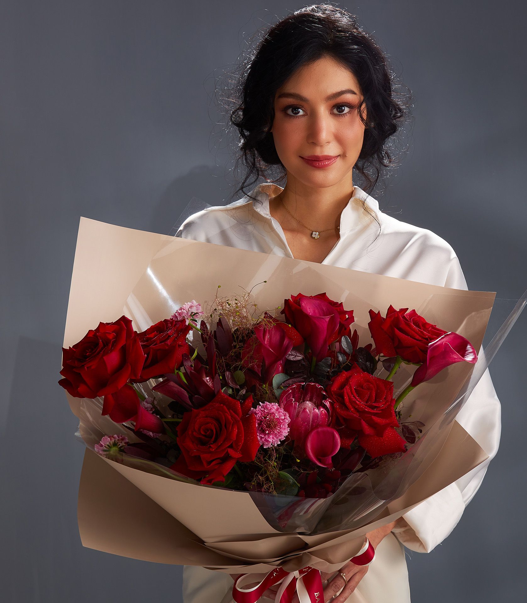 Woman holding large red rose and pink calla lily bouquet with greenery, wrapped in kraft paper and red ribbon