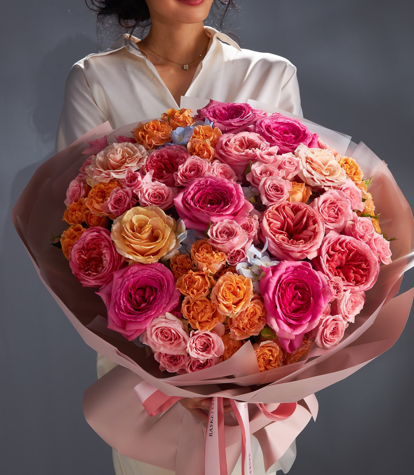 Large luxury rose bouquet in pink, fuchsia, and peach with blue accents, wrapped in pastel paper with pink ribbons for