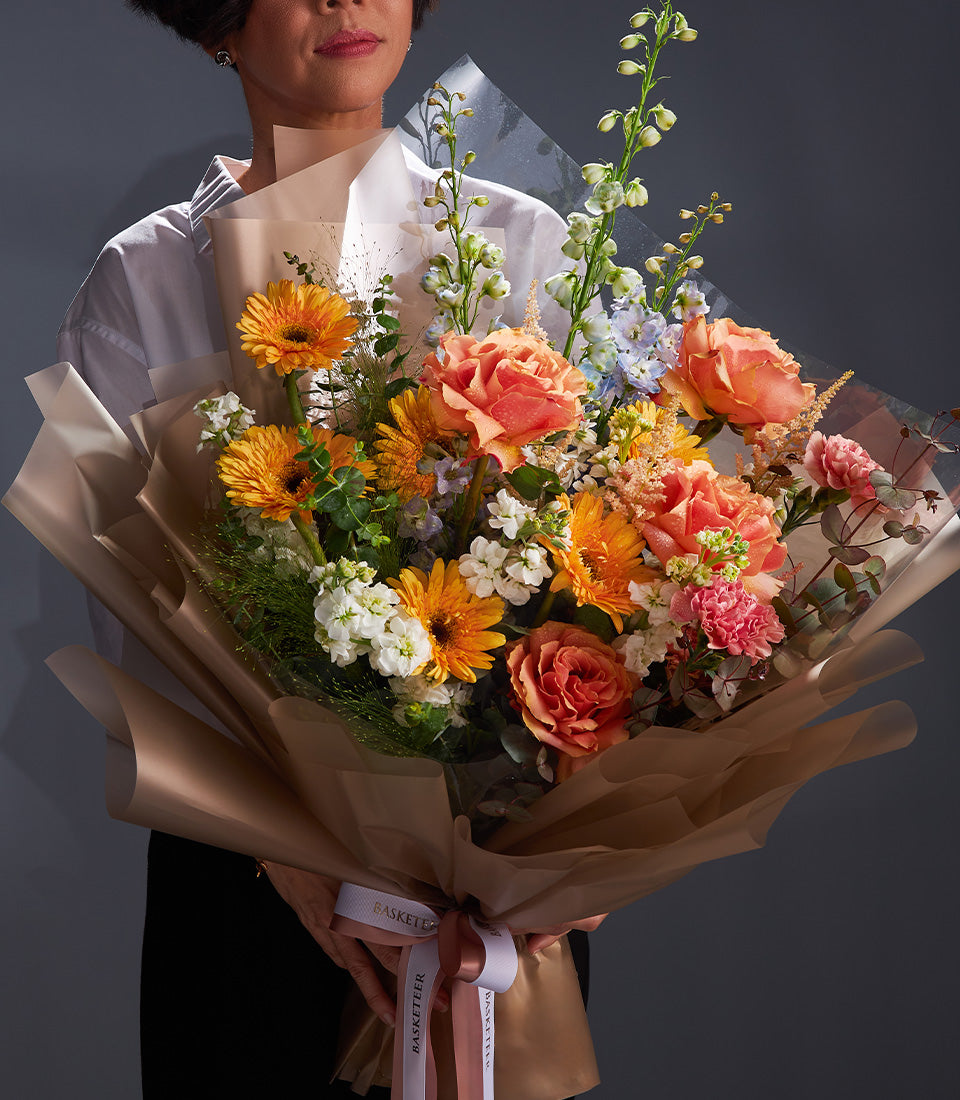 Vibrant mixed flower bouquet with orange gerberas, peach roses, and blue delphiniums, wrapped in kraft paper