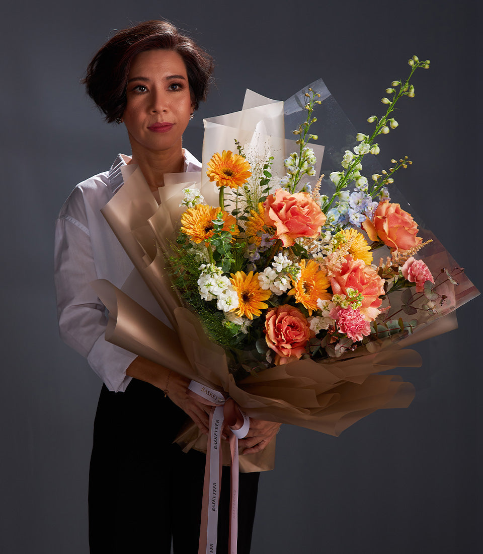 Vibrant bouquet of orange gerbera daisies, coral roses, and white stock flowers wrapped in brown paper with white ribbon