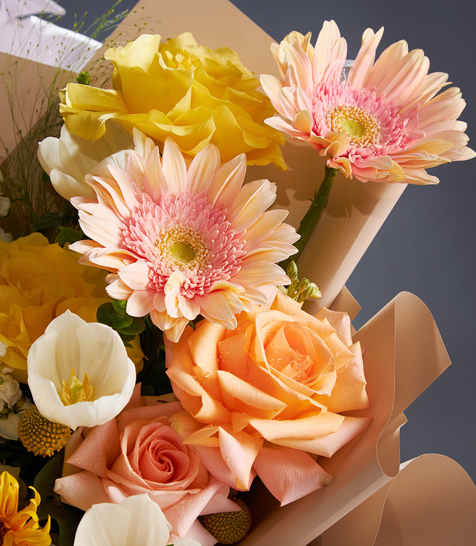 Vibrant mixed flower bouquet with yellow roses, pink gerbera daisies, peach roses, and white tulips in beige paper wrap