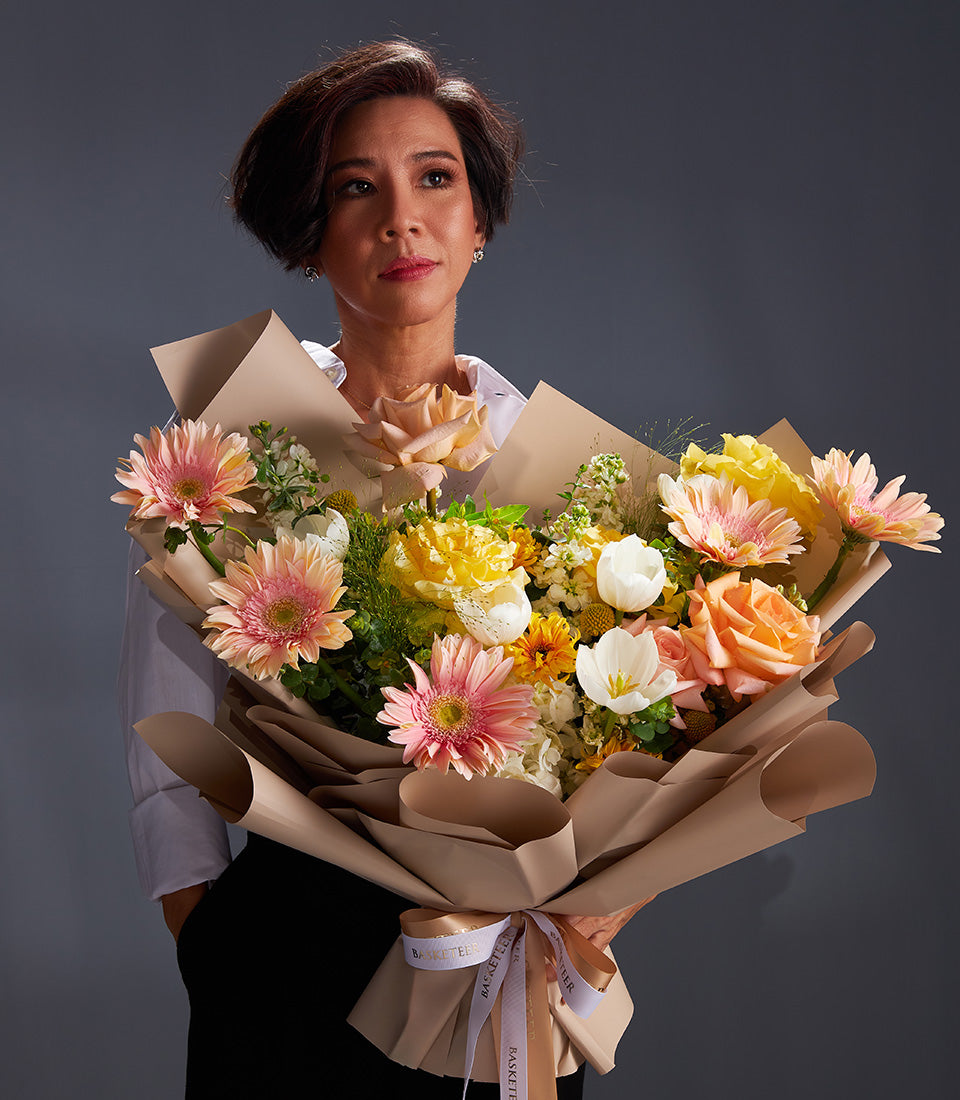 Woman holding vibrant mixed flower bouquet with pink roses, yellow gerbera daisies, and orange tulips in brown paper wrap