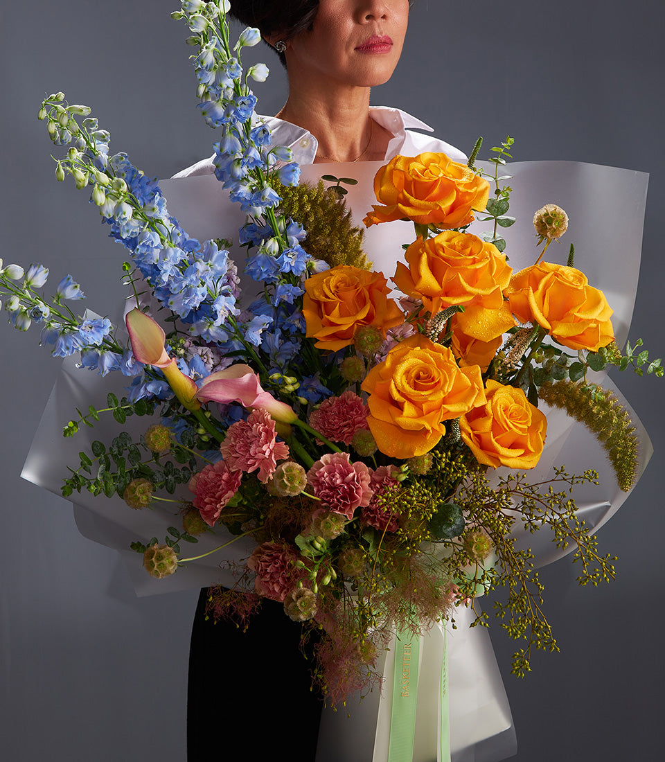 Premium mixed bouquet with orange roses, blue delphiniums, and pink carnations in white wrapping