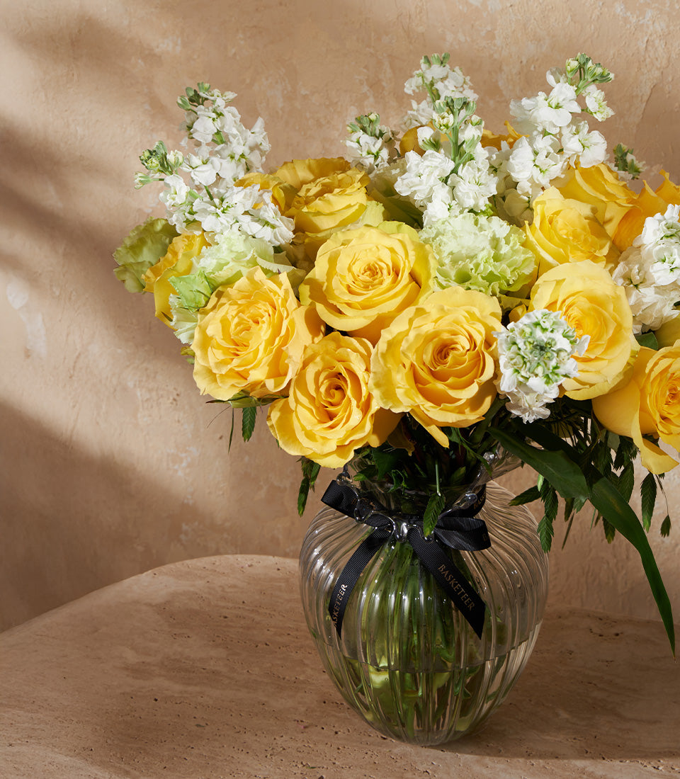Bright yellow roses with white filler flowers in a ribbed glass vase, tied with black ribbon