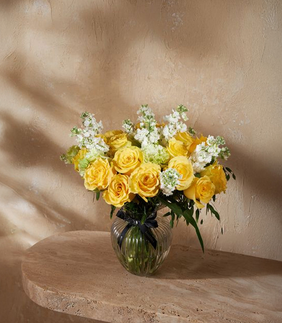 Yellow roses and white stock flowers in a clear glass vase with black ribbon, fresh flower gift arrangement