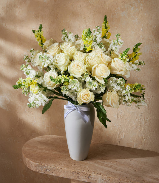 Elegant white vase arrangement with ivory roses, yellow snapdragons, and green filler flowers