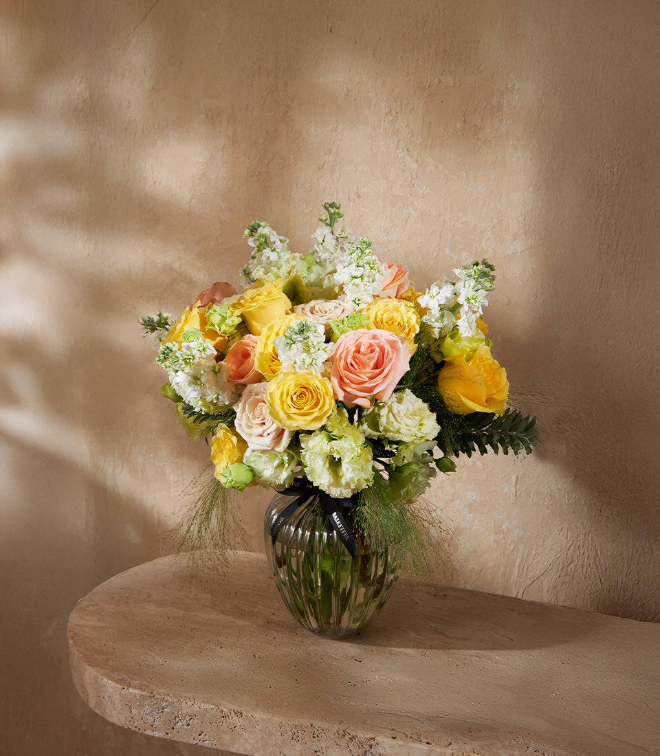 Yellow, peach and pink rose vase arrangement with stock flowers and green foliage, tied with black ribbon