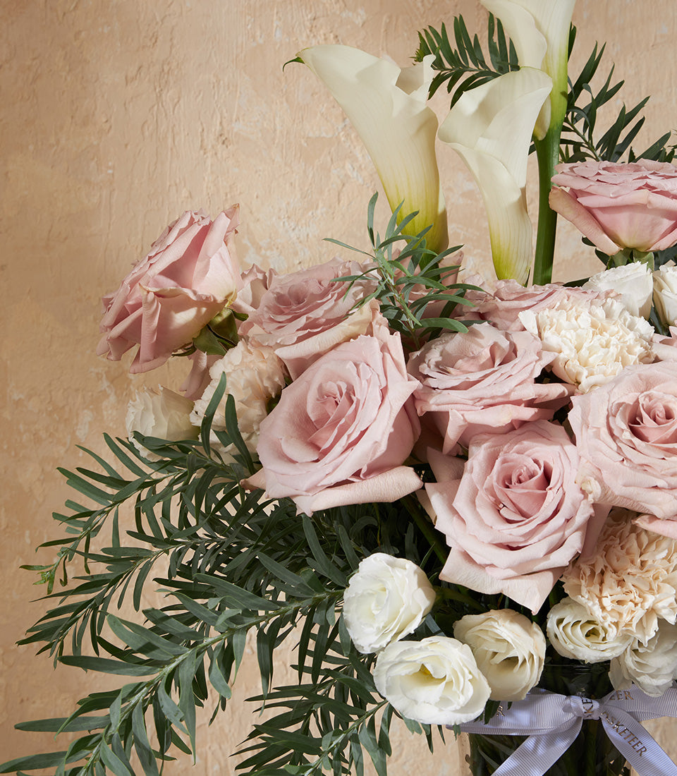 Pale pink roses, white calla lilies, and peach carnations in clear glass vase with grey ribbon