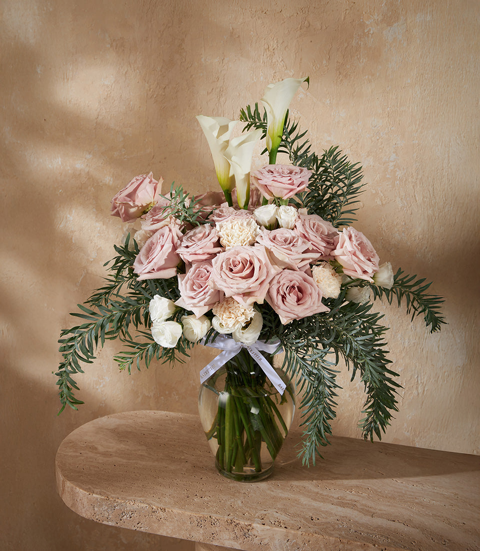 Dusty pink roses with white calla lilies and carnations in clear glass vase with green foliage