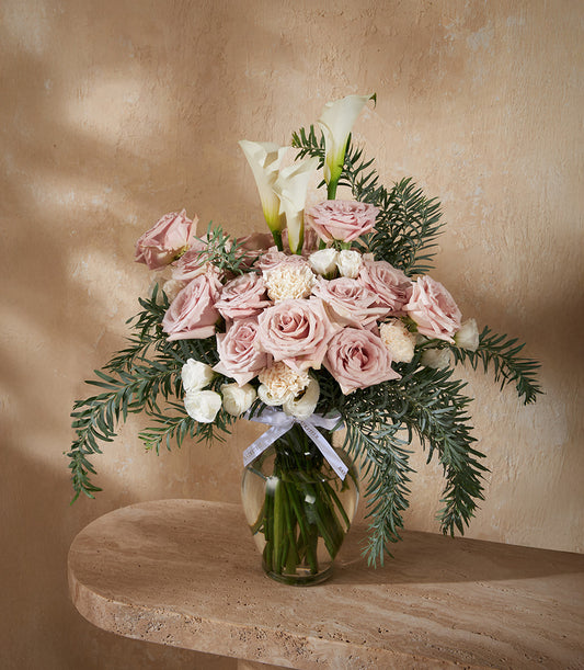 Dusty pink roses with white calla lilies and carnations in clear glass vase with green foliage