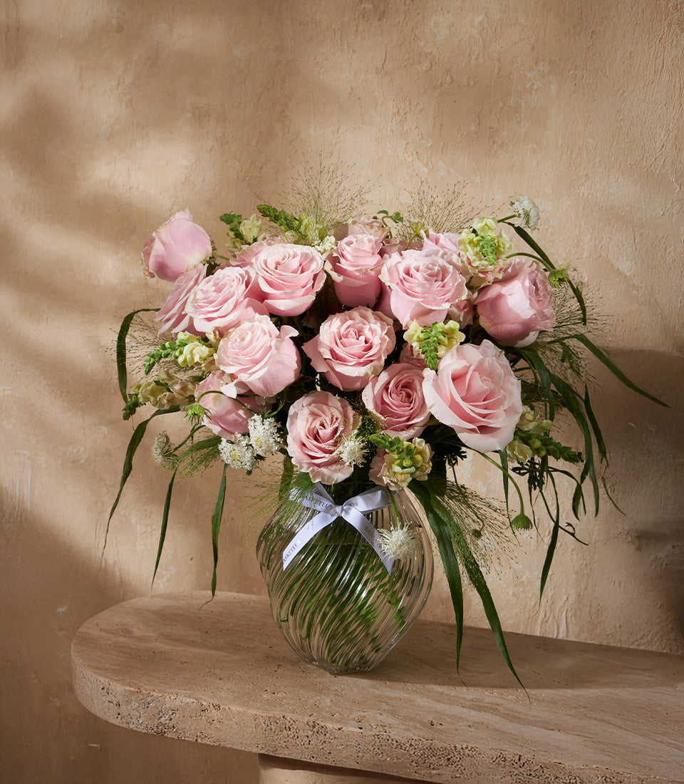 Soft pink roses with mixed greenery in clear ribbed glass vase, white ribbon accent