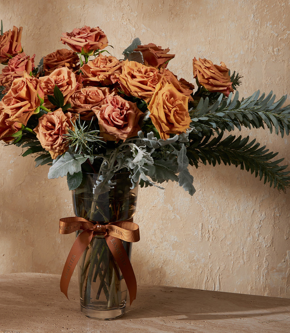 Vibrant orange and rust roses in clear glass vase with green foliage and brown satin ribbon bow