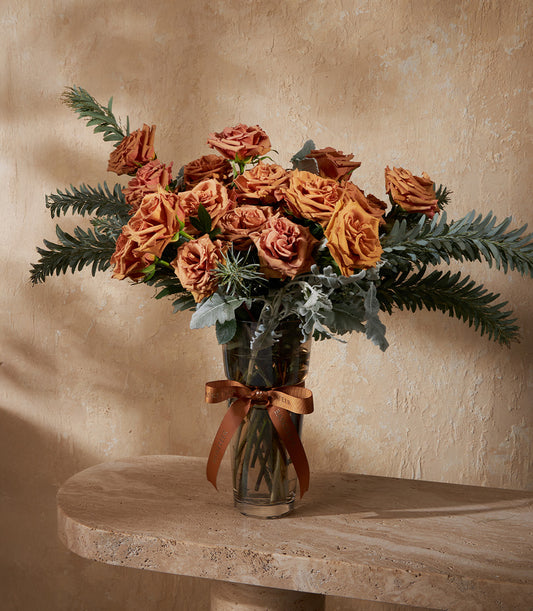 Burnt orange and terracotta rose arrangement in clear glass vase with brown satin ribbon