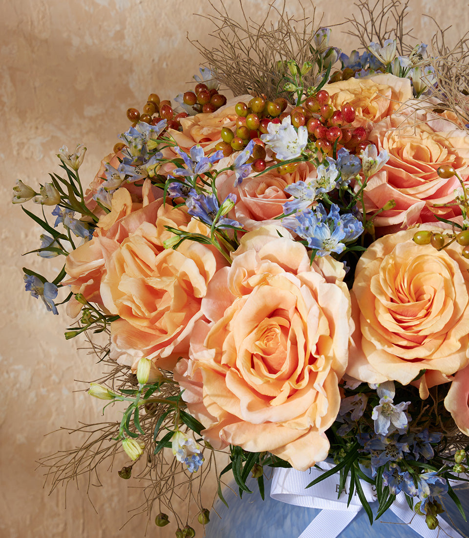 Peach and orange roses with delphiniums and berries in light blue vase, tied with white ribbon