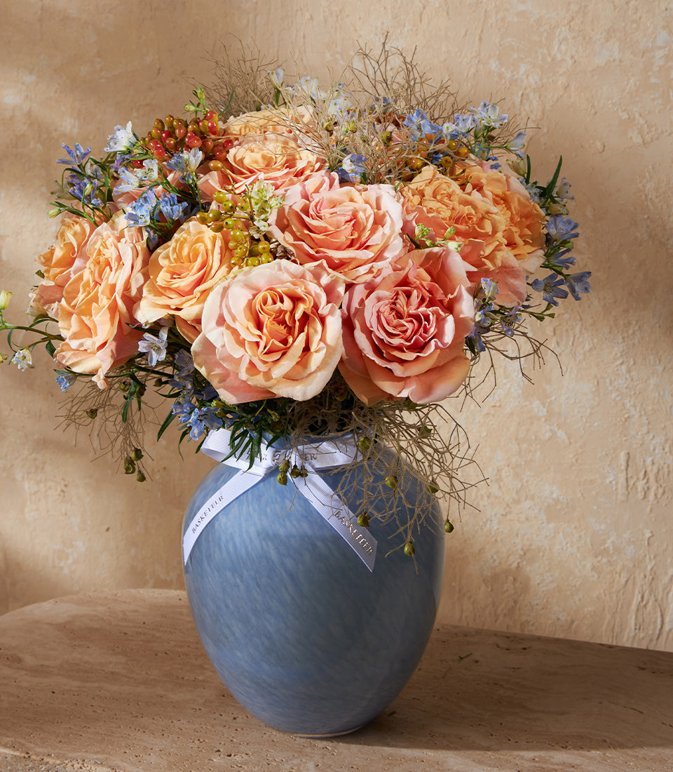 Peach and coral roses with blue accents in swirled blue glass vase, premium fresh flower arrangement