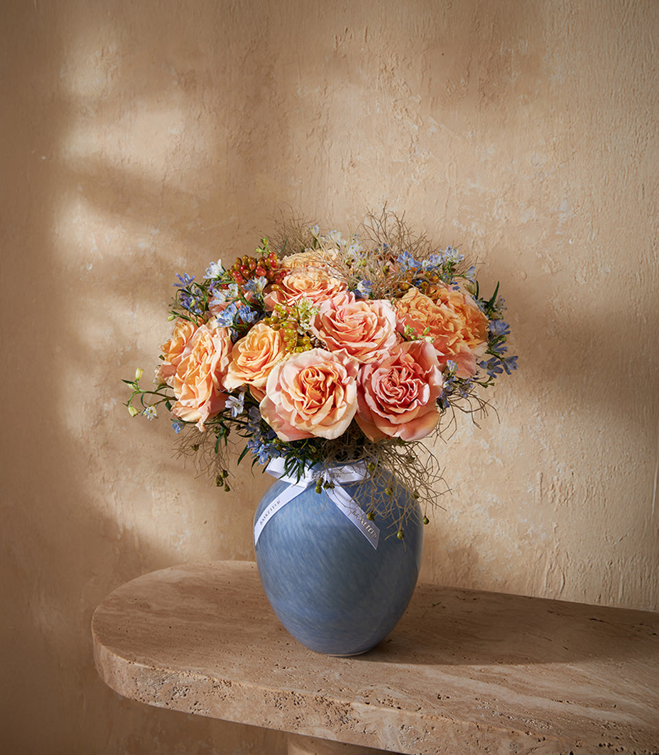 Peach and coral roses with blue flowers in marbled glass vase, tied with white ribbon