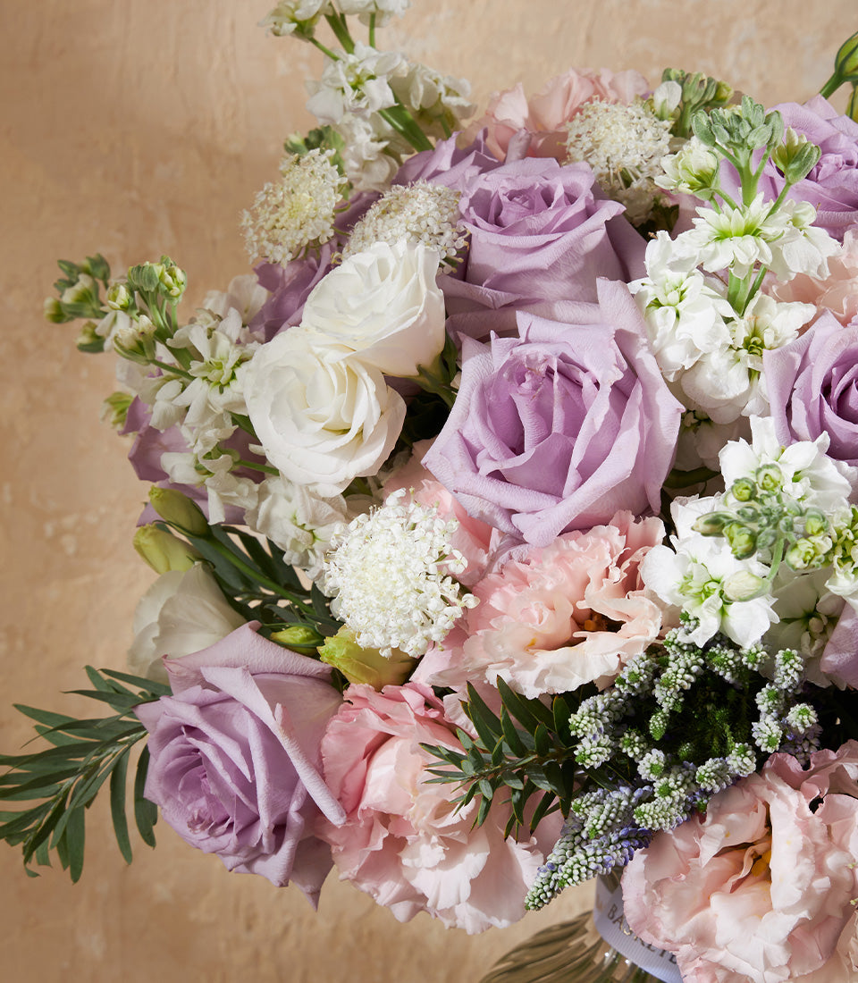 Lavender roses, pink carnations, and white blooms in a clear glass vase with soft green foliage
