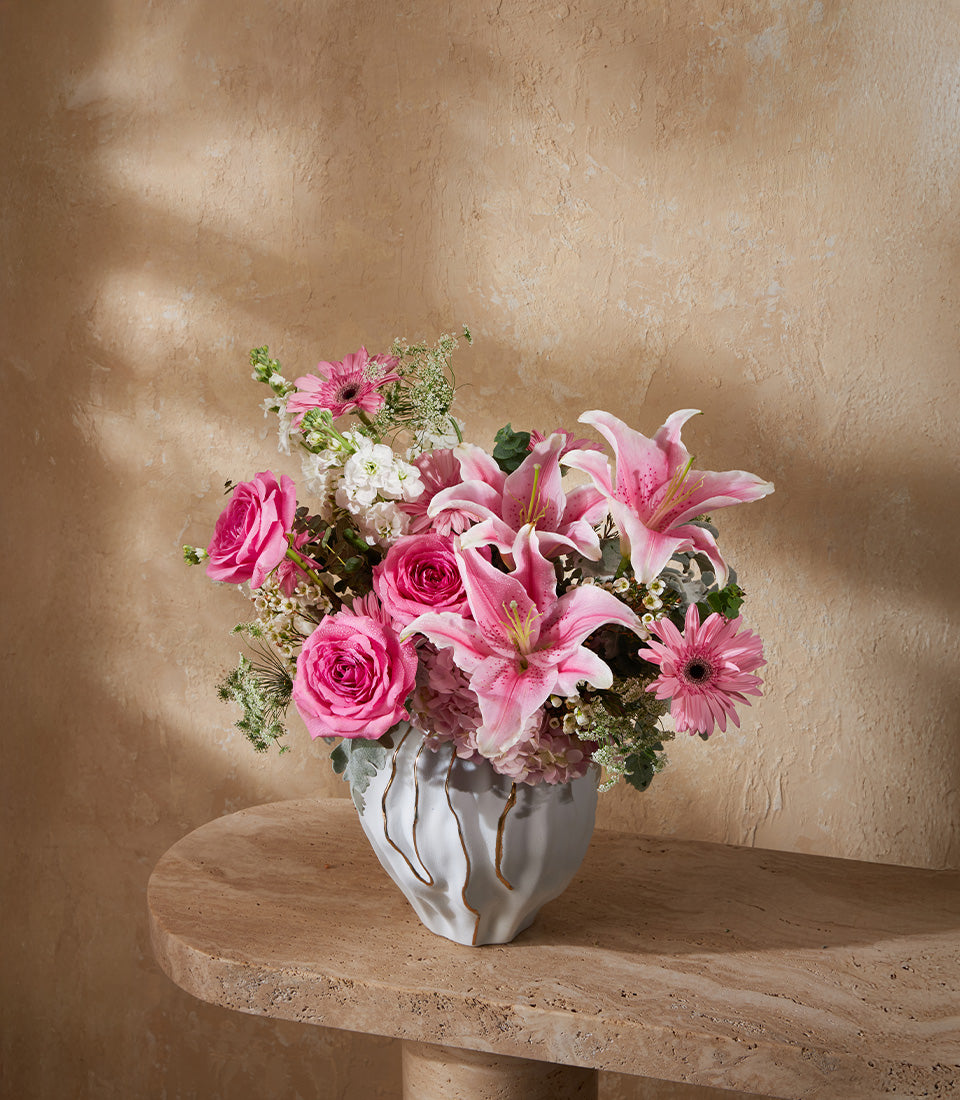 Pink and white lilies, roses, and gerberas in textured grey ceramic vase with gold accents