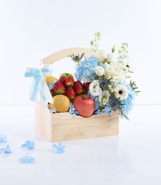 Wooden gift basket with fresh strawberries, apples, citrus fruits, white roses, and blue hydrangeas for Mother's Day